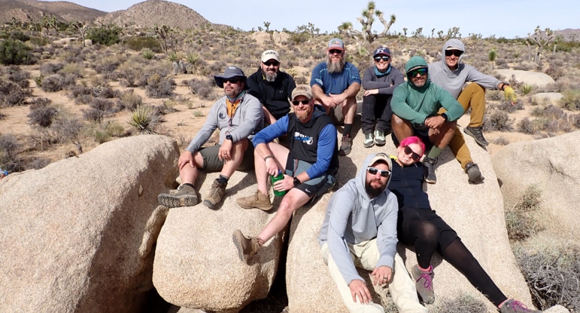 a group of veterans pose for a group photo while sitting on a large rock formation in joshua tree